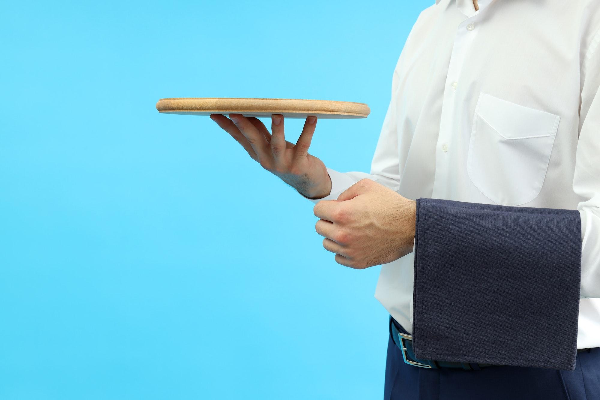 Young man waiter holds tray on blue background, space for text