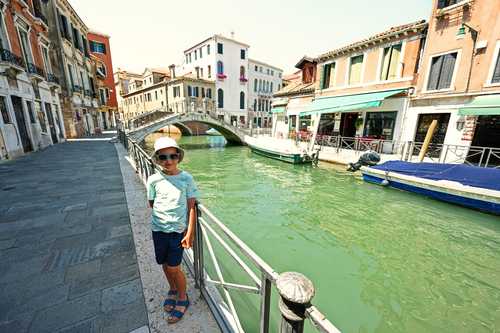 Boy in panama and sunglasses stand near canal in Venice, Italy.