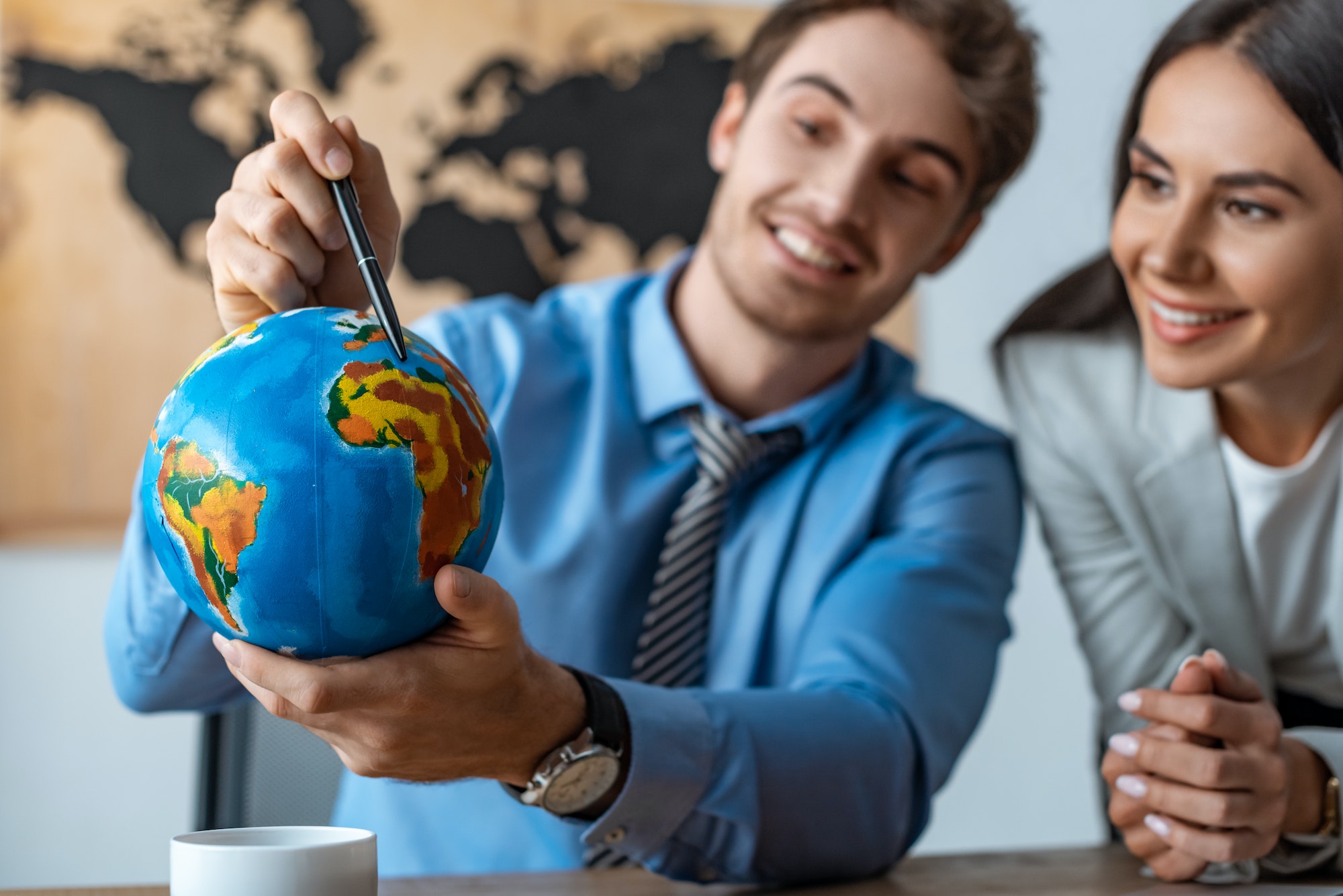 selective focus of attractive travel agent standing near smiling colleague pointing with pen at