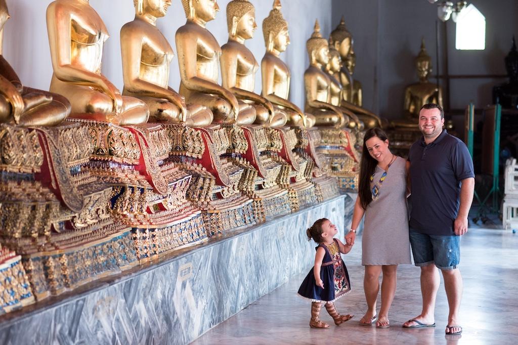 A mother and father holding hands with their young daughter while walking past a long row of golden seated Buddha statues in a Thai temple, capturing a moment of educational family travel.