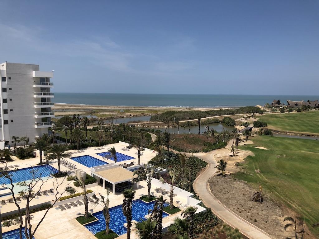 An aerial view of a sprawling beachfront resort in Cartagena featuring multiple blue swimming pools, a green golf course, and a white modern hotel building, offering a variety of amenities for family travel.