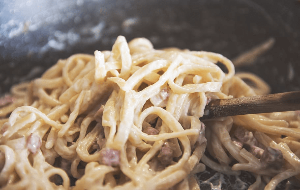 A close-up shot of a wooden spoon tossing creamy Spaghetti alla Carbonara with pancetta in a pan, capturing the glossy emulsion of the sauce.
