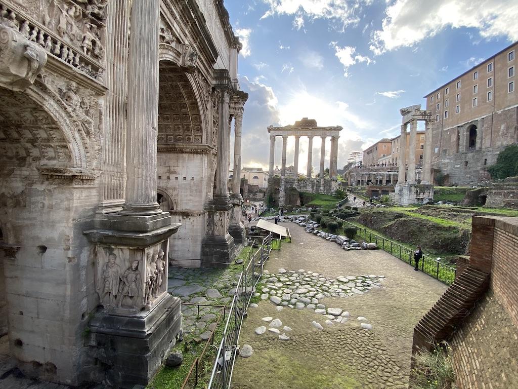 A detailed view of the Arch of Septimius Severus featuring intricate stone reliefs, with the columns of the Temple of Saturn in the distance at the Roman Forum, a must-see for those who travel Rome.