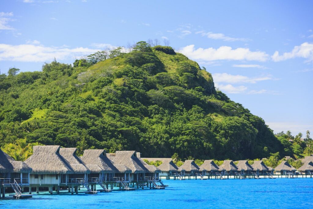 Traditional thatched-roof overwater bungalows sit above a turquoise lagoon, framed by lush tropical greenery at a luxury Bora Bora resort in French Polynesia.