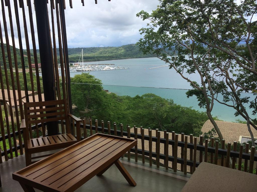 A wooden patio table and chair on a balcony overlooking a lush green coastline and a harbor with white sailboats, a peaceful view for those who travel Costa Rica.