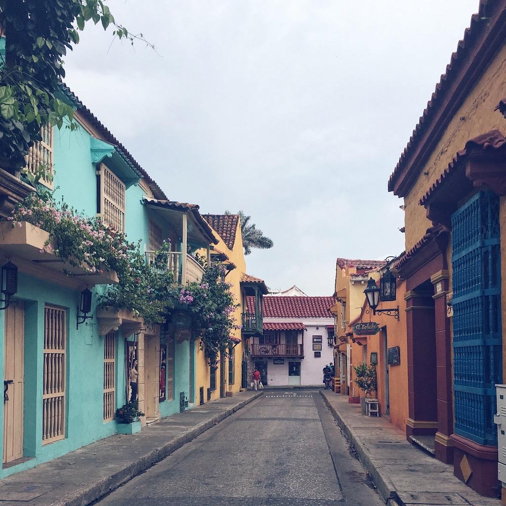 A view down a narrow, cobblestone street lined with colorful colonial buildings featuring wooden balconies and blooming bougainvillea in the city of Cartagena.