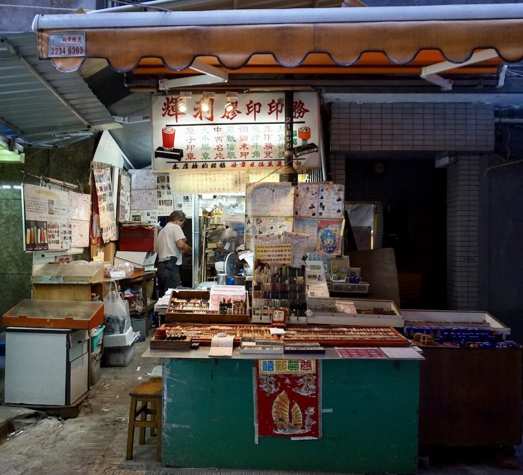 A street-level view of a traditional artisan stall in Sheung Wan's Man Wa Lane, also known as "Chop Alley," where skilled craftsmen hand-carve custom Chinese seals and stamps for those who travel Hong Kong.