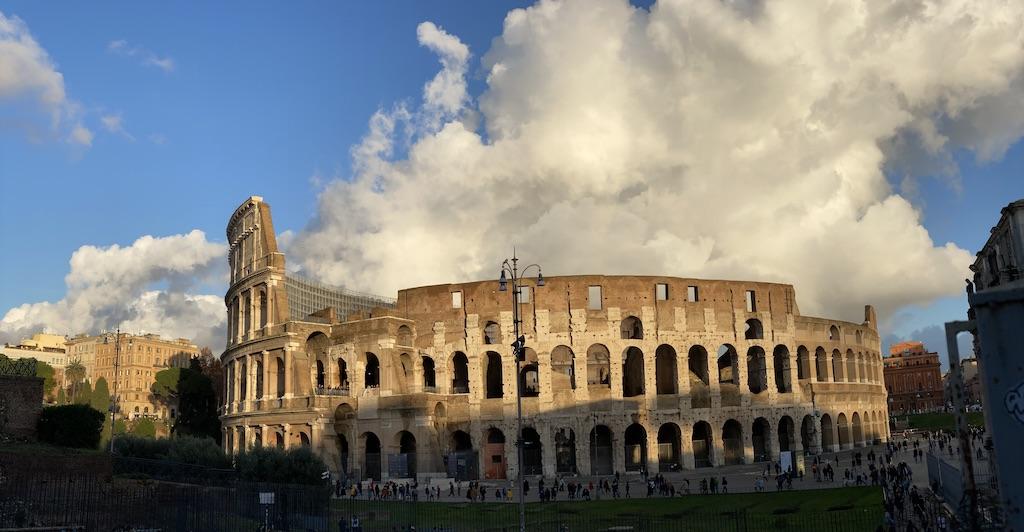 A side-angle view of the ancient stone tiers and arches of the Colosseum amphitheater under a dramatic cloudy blue sky, a centerpiece for any travel Rome itinerary.