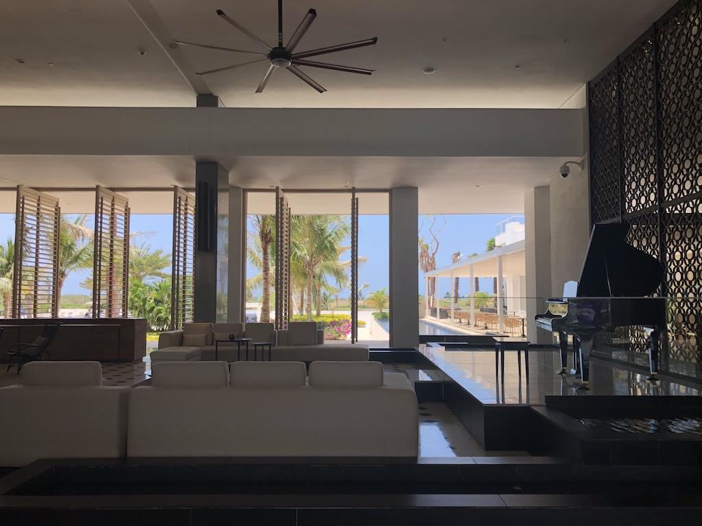 A view from a minimalist hotel lobby featuring white modular sofas and a black grand piano, looking out toward a tropical poolside terrace through floor-to-ceiling windows for those who travel Cartagena.