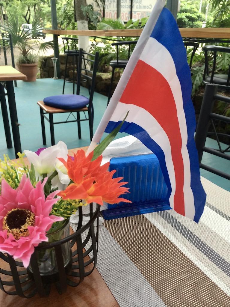 The national flag of Costa Rica stands in a small holder next to a basket of pink and orange flowers on a patio table, welcoming those who travel Costa Rica.