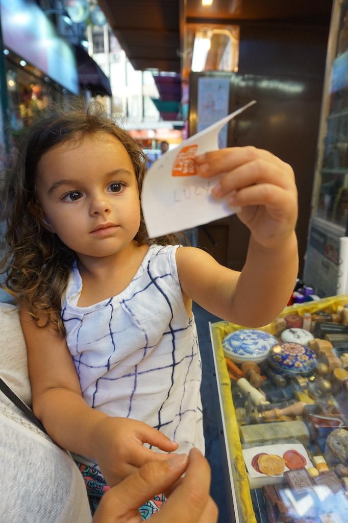 A local artisan in a green apron at a traditional street stall in Sheung Wan's Man Wa Lane, carefully showing a young girl how to use a hand-carved Chinese seal, a popular souvenir for those who travel Hong Kong.