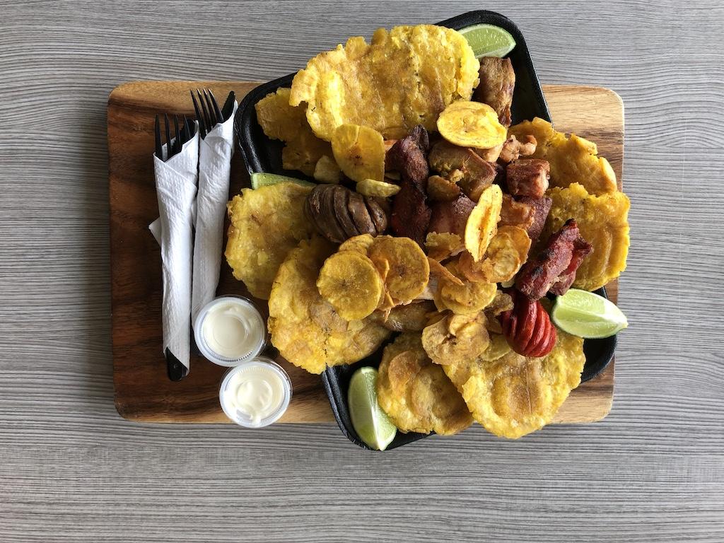 A top-down view of a wooden board holding a platter of crispy fried plantains, grilled meats, and lime wedges, offering a flavorful alternative to standard pita food.