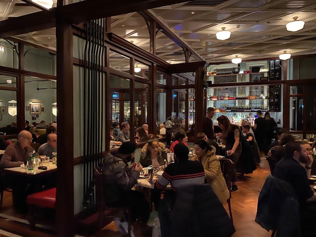 Patrons dining inside a warm, wood-paneled brasserie with high ceilings and an illuminated back bar, a popular culinary stop for those who travel Manchester.