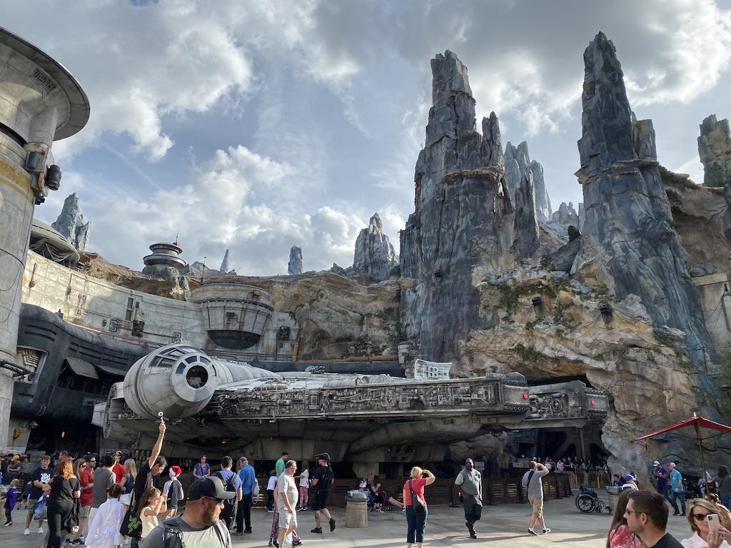 A crowd of visitors, including many children, walking past a life-sized Millennium Falcon spaceship replica and jagged spires at a popular theme park, illustrating an immersive destination for family travel.