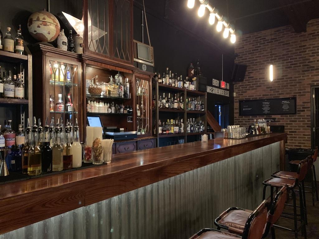 An empty long wooden bar at a dimly lit industrial-style pub with brick walls and shelves of spirits, a popular nightlife spot for those who travel Manchester.