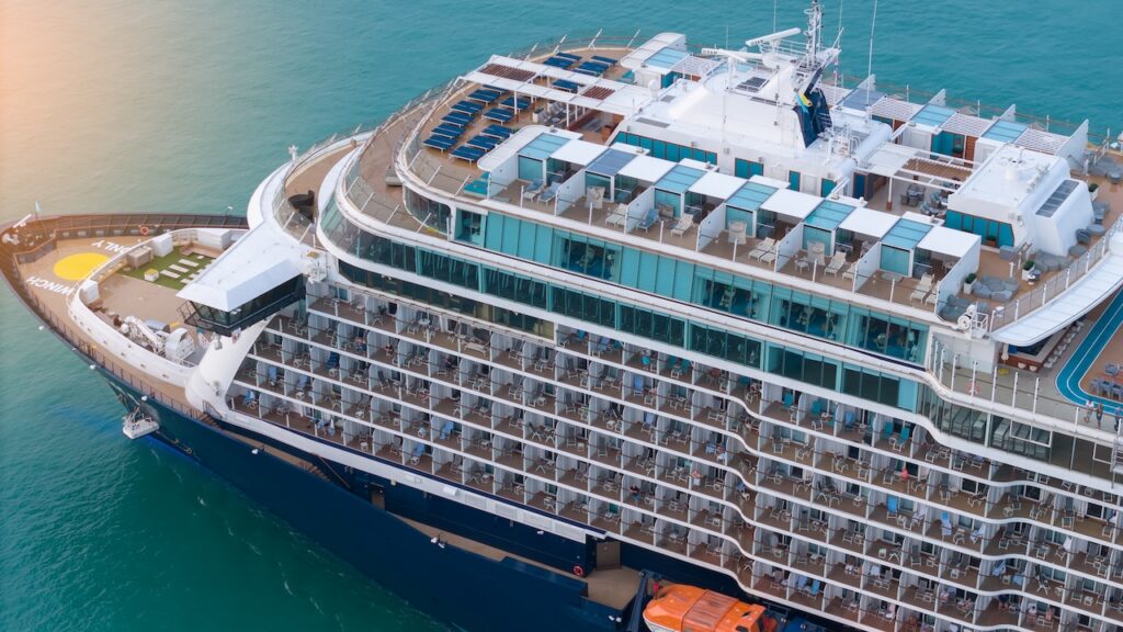 High-angle aerial view of a luxury cruise ship's upper decks, showing numerous private passenger balconies, tinted windows, and open sundeck areas overlooking the turquoise ocean.