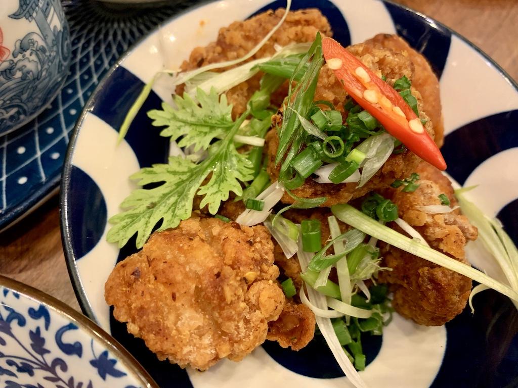 Top-down view of golden-brown fried chicken topped with sliced red chili, fresh cilantro, and spring onions on a blue patterned plate, representing authentic street food found when you travel Bangkok Thailand.