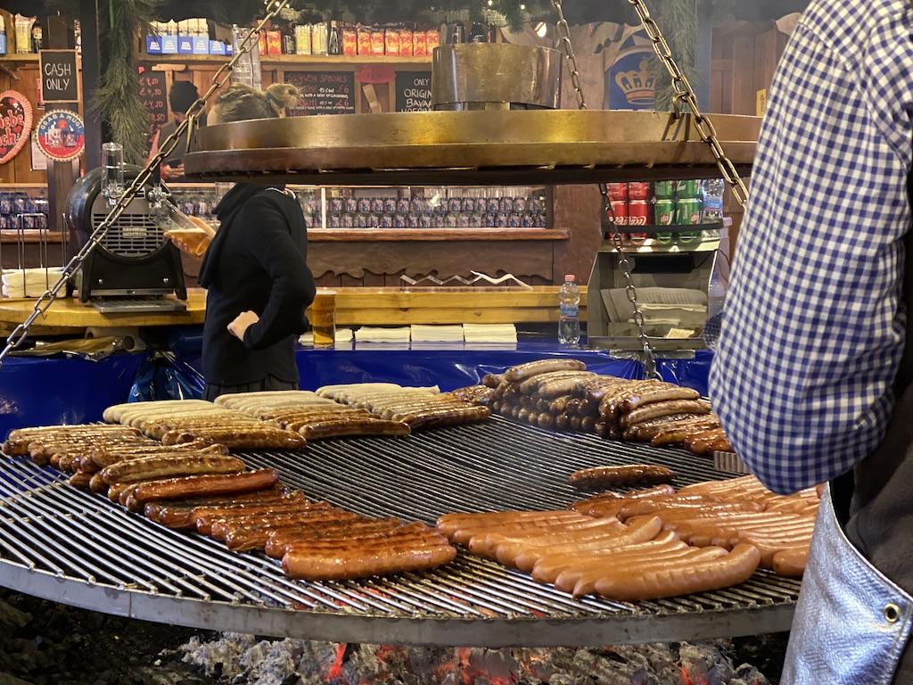 Dozens of sausages and bratwursts cooking on a large circular swinging grill at an outdoor food stall, a staple experience for those who travel Manchester during the holiday season.