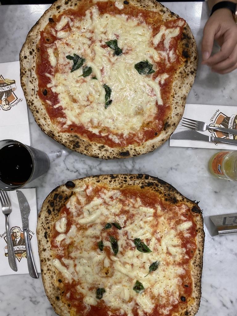 Top-down view of two large, thin-crust Neapolitan pizzas with melted mozzarella and fresh basil on a marble table at a pizzeria, an essential meal when you travel Rome.