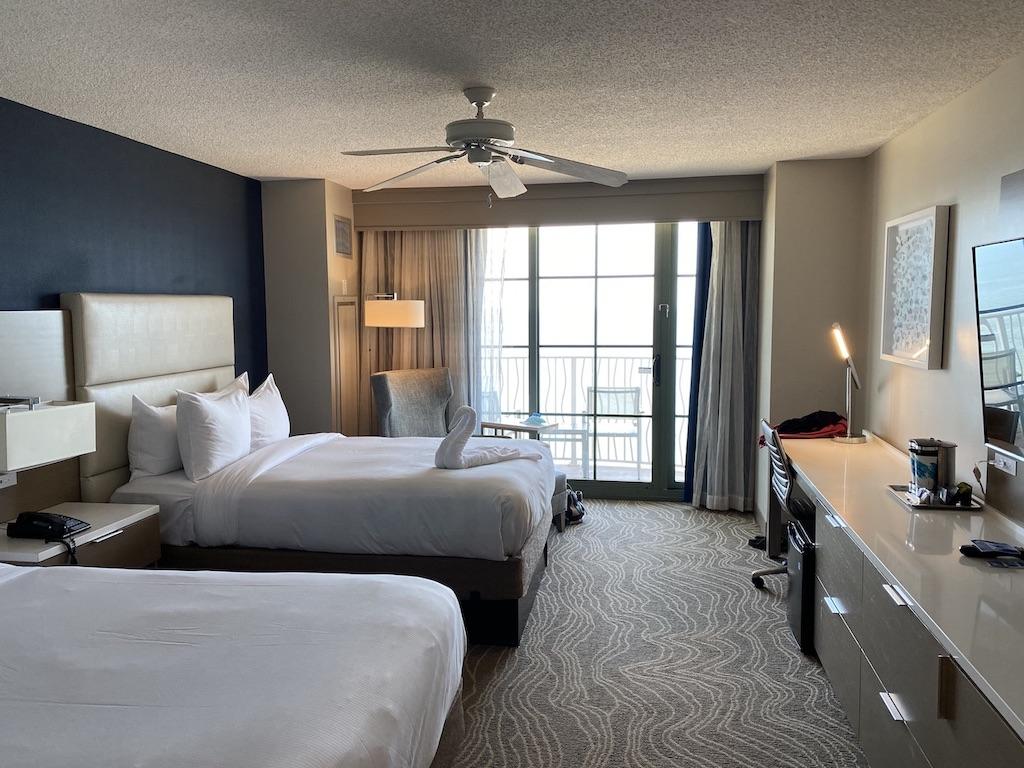A hotel room in Virginia Beach featuring two beds with white linens, a large window leading to a balcony, and a patterned carpet.