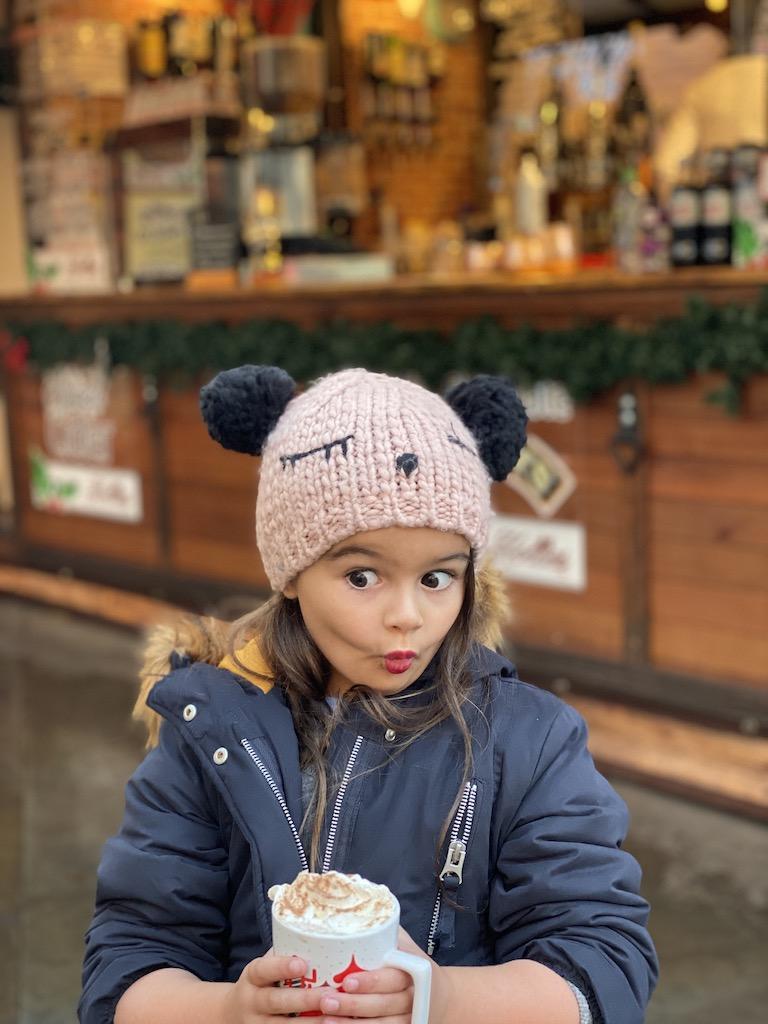 A young girl in a pink knit pom-pom hat making a playful face while holding a cup of whipped cream-topped cocoa at a Christmas market, a festive highlight for those who travel Manchester.
