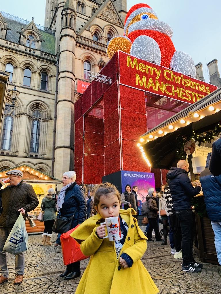 A young girl in a bright yellow coat sips a drink in front of the festive stalls and large "Merry Christmas Manchester" sign at the Manchester Christmas markets.