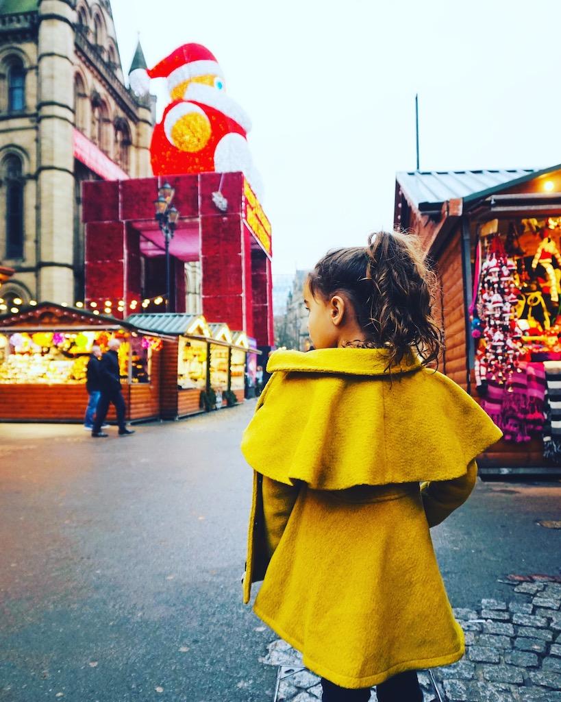 A young girl in a yellow coat looks toward festive wooden stalls and a large red holiday structure at the Manchester Christmas markets.
