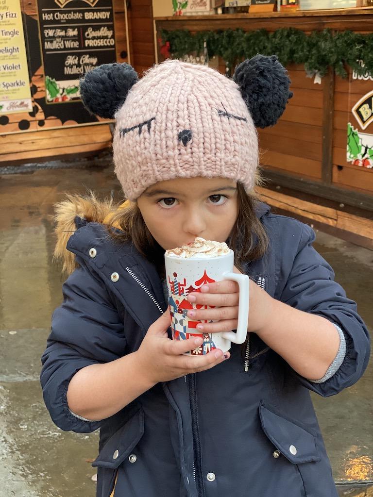 A young girl in a pink knitted hat with pom-poms drinks a mug of hot chocolate topped with whipped cream, a cozy scene reminiscent of the festive spirit found at Manchester Christmas markets.