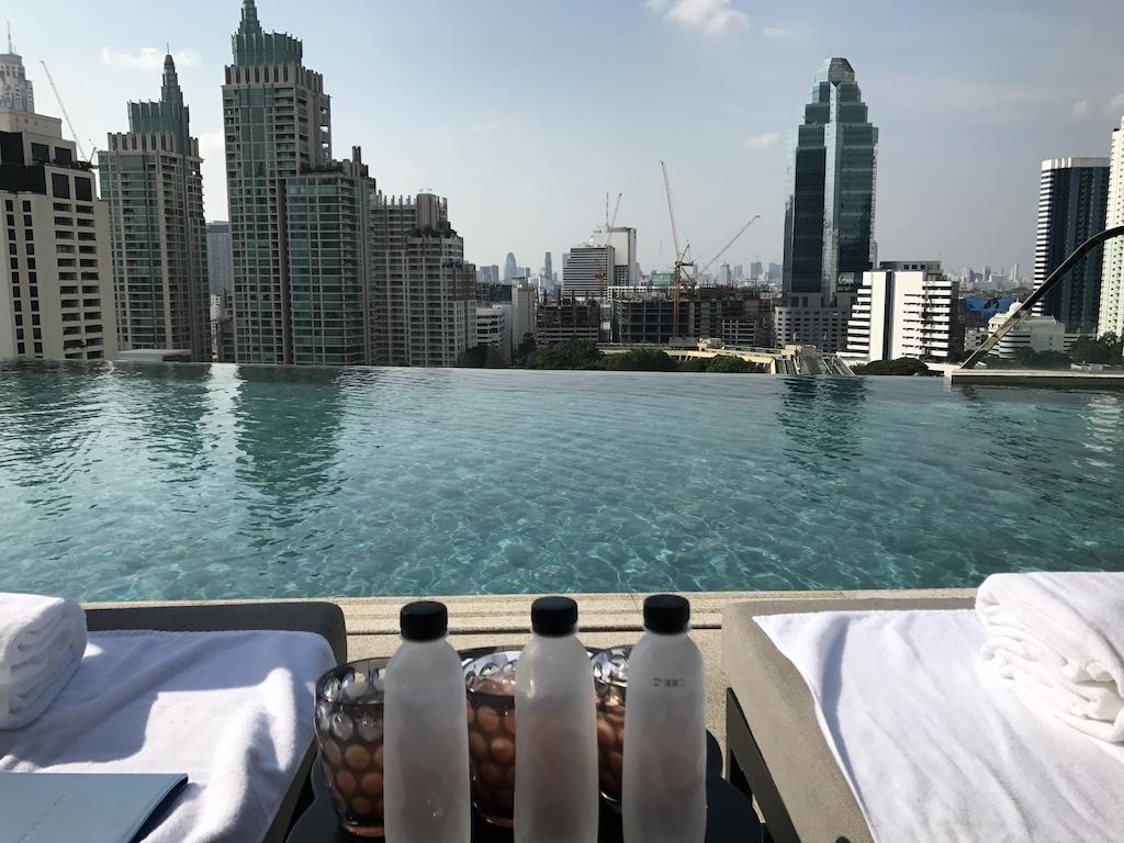 Rooftop infinity pool overlooking a modern city skyline with skyscrapers and construction cranes, featuring lounge chairs and chilled water bottles for those who travel Bangkok Thailand.