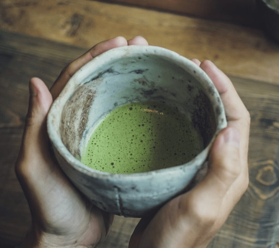 Hands cupping a rustic stone bowl filled with frothy green matcha tea, capturing a traditional ceremony inside a japanese tea house.
