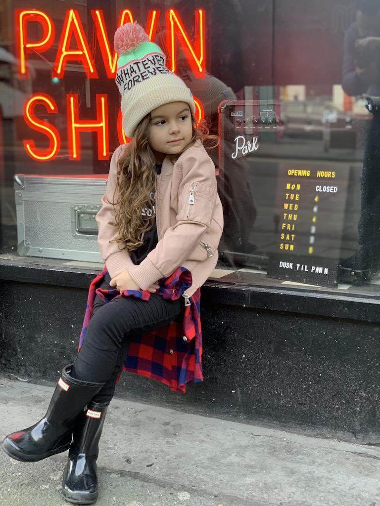 A young girl in a pink bomber jacket, beanie, and rain boots sits outside a storefront with a red neon "Pawn Shop" sign and "Dusk til Pawn" hours, showcasing the edgy city vibe for those who travel Manchester.