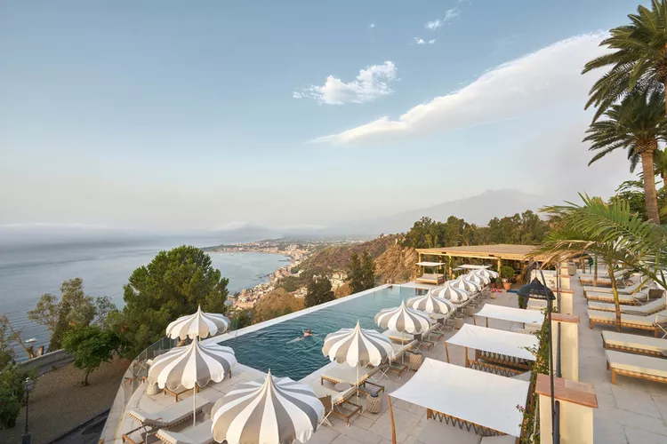 A scenic view of the White Lotus Hotel featuring an outdoor pool area with white sun loungers and umbrellas, overlooking a body of water and lush green landscape under a partly cloudy sky.