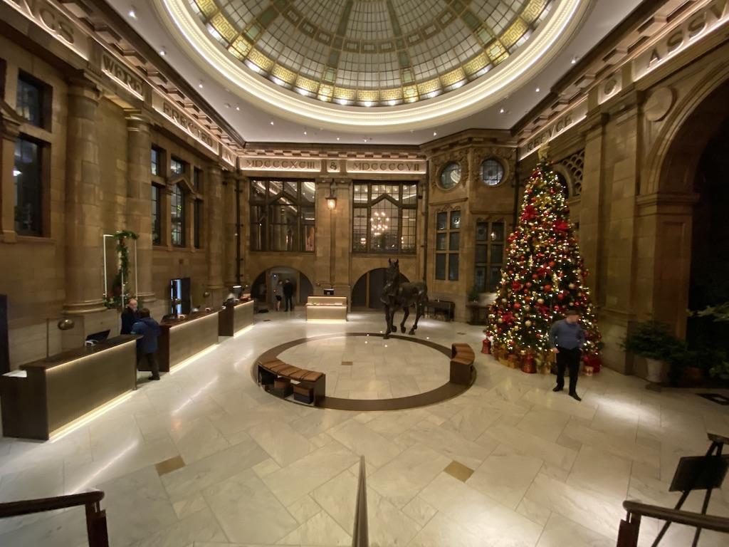 A grand circular hotel lobby featuring a domed stained-glass ceiling, stone pillars, and a towering red and gold Christmas tree, a luxurious stay for those who travel Manchester.