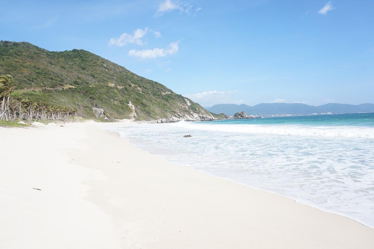 A wide shot of a pristine white sand beach meeting turquoise ocean waves under a bright blue sky, with lush green hills in the background, perfect for travelers applying experience to find secluded coastal gems.