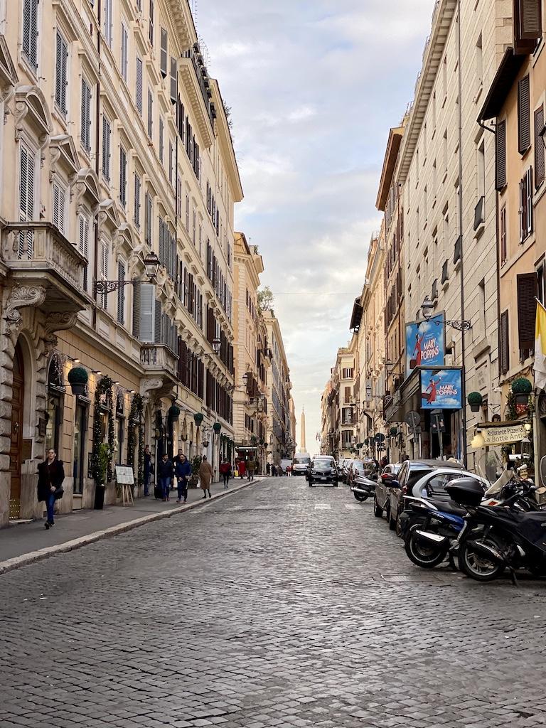 A long cobble-stoned street lined with classic multi-story Italian buildings, parked scooters, and a distant view of an obelisk under a soft sky, perfect for a travel rome exploration.