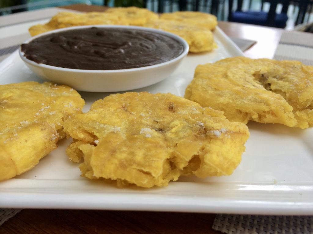 A close-up of crispy, salted patacones served with a side of smooth black bean dip on a white rectangular plate, a popular appetizer to try when you travel Costa Rica.