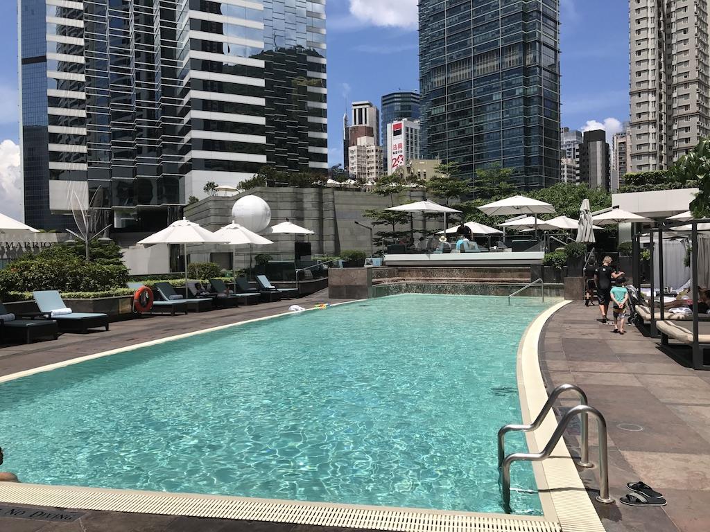 A sparkling outdoor swimming pool surrounded by lounge chairs and white umbrellas, set against a backdrop of towering skyscrapers for a luxury travel Hong Kong experience.