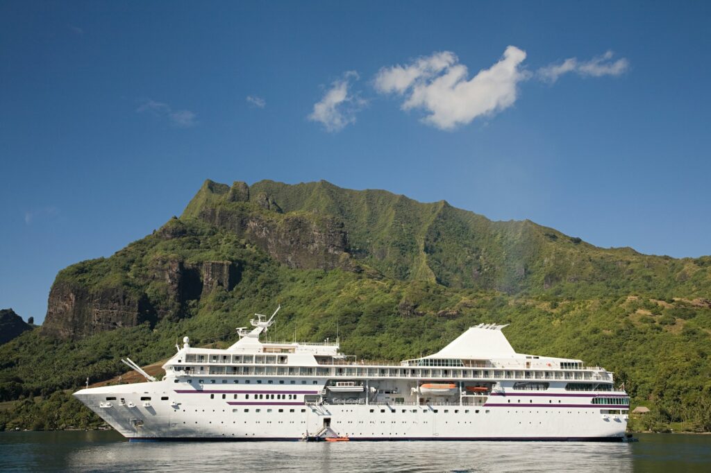 A large, white cruise ship with purple trim is anchored on calm water below a massive, lush, green mountain covered in dense tropical foliage, all set against a clear blue sky with a single white cloud.
