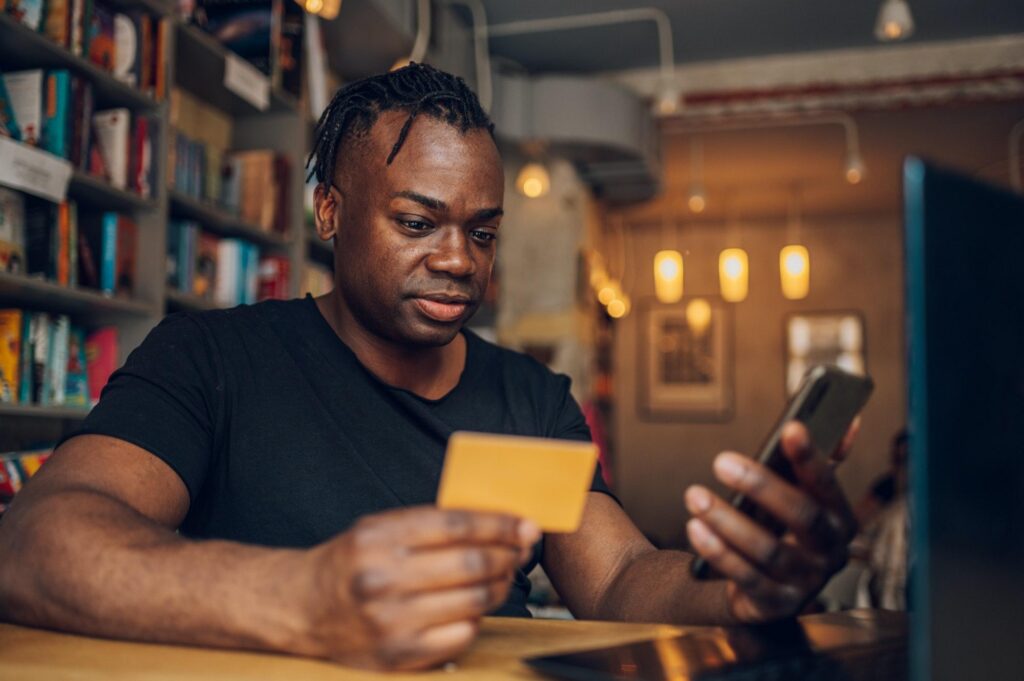 A man sits in a bookstore or cafe, holding a yellow business credit card while looking at his smartphone to manage expenses or complete a purchase.