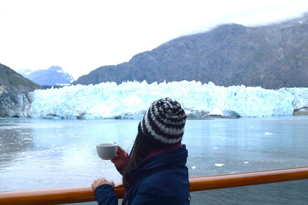 Woman in a winter hat and jacket holding a warm cup, viewing a massive blue glacier and icy mountain range from the railing of an Alaska cruise ship on a cold, overcast day.
