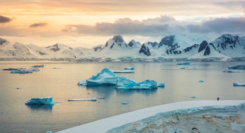 Tourists in a zodiac boat navigate through a field of massive icebergs under a golden sky during an Antarctic cruise.