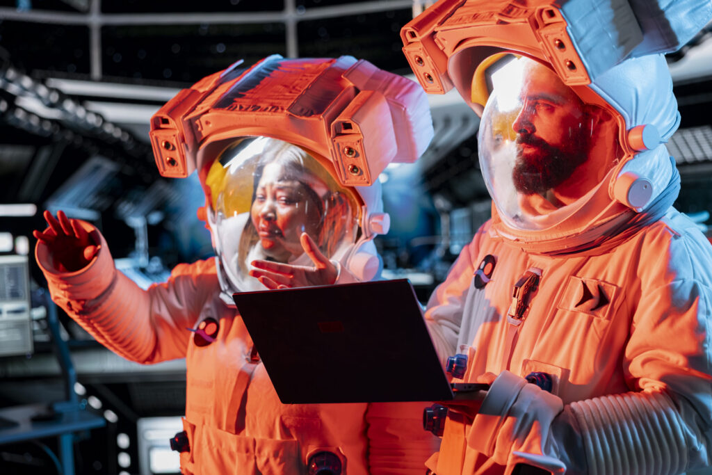 Two astronauts in glowing helmets work on a laptop inside a futuristic cabin, symbolizing the rise of space tourism industry.