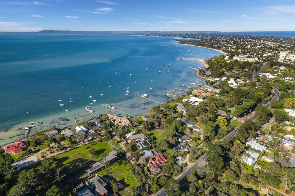 Aerial view of a picturesque coastal town with beachfront homes and a deep blue bay filled with sailboats, emphasizing luxury seaside living and scenic travel.