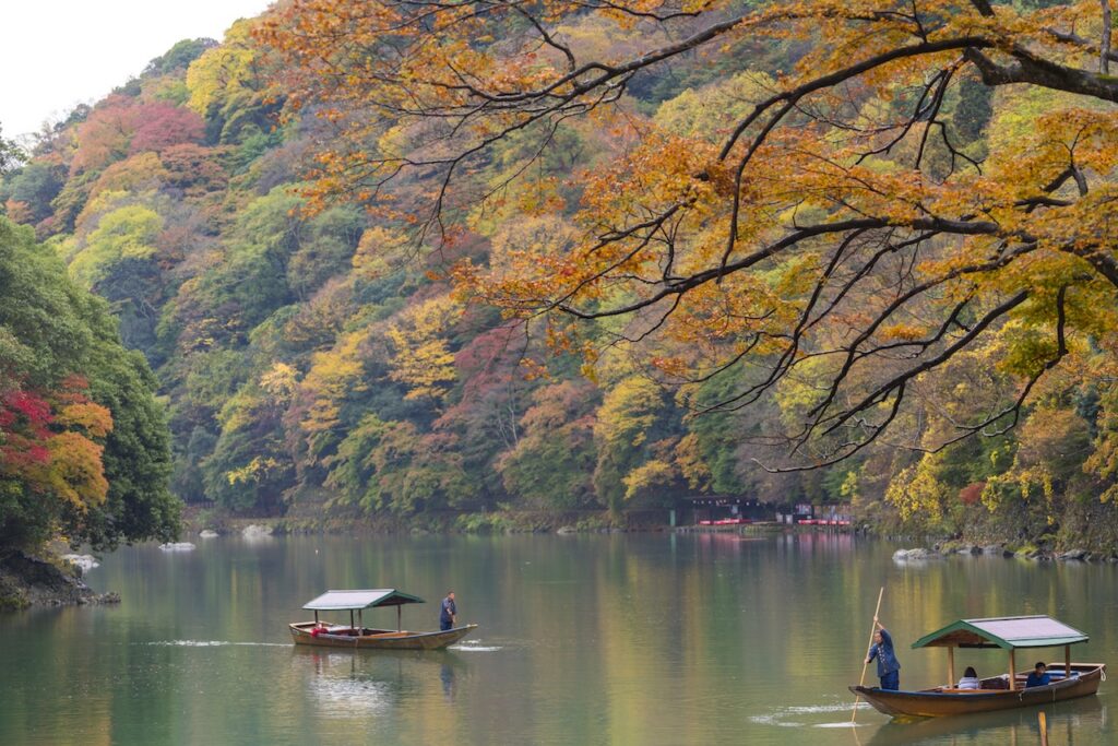 View of a river or lake flanked by a steep hill covered in autumn foliage, primarily yellow, orange, and green. Two small, traditional wooden boats are floating on the water.