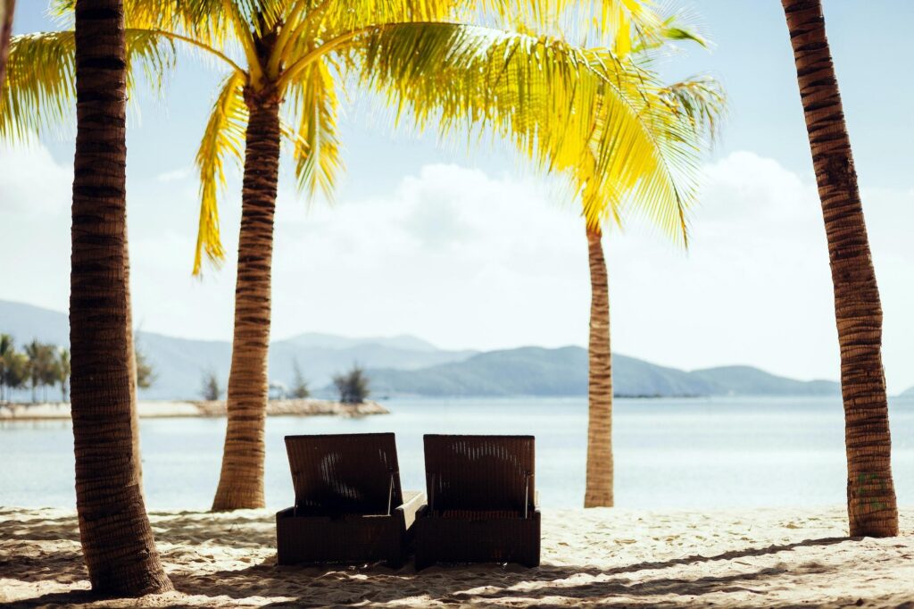 Two lounge chairs shaded by palm trees overlook a calm bay and the mountainous landscape of the Hawaiian islands.