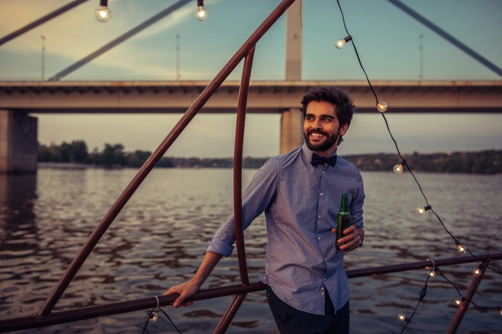 A smiling man in a blue button-down shirt and bowtie holds a green bottle of beer while leaning against a railing at sunset, with a large bridge and calm river in the background. This relaxed evening scene represents the upscale social life enjoyed by virgin adults on a luxury adults only cruise.