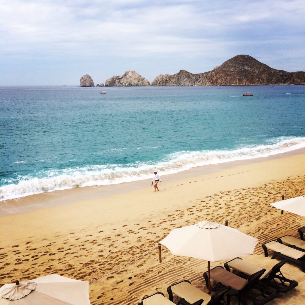 A rugged coastal view of a secluded sandy beach near Rancho Pescadero Cabo, featuring dark rocky cliffs where seagulls are perched. The gentle waves of the Pacific Ocean meet the shoreline under a soft, overcast sky, highlighting the natural beauty of the Baja California Sur region.