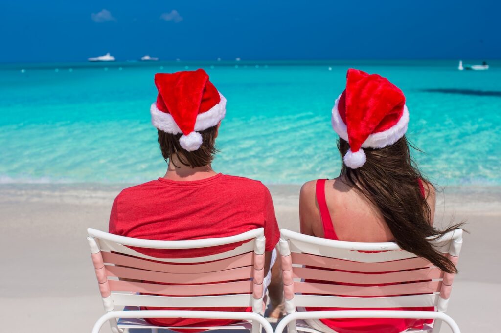 Couple in Santa hats relaxing on a tropical beach with turquoise water, representing a warm-weather Silversea Christmas cruise in 2025.