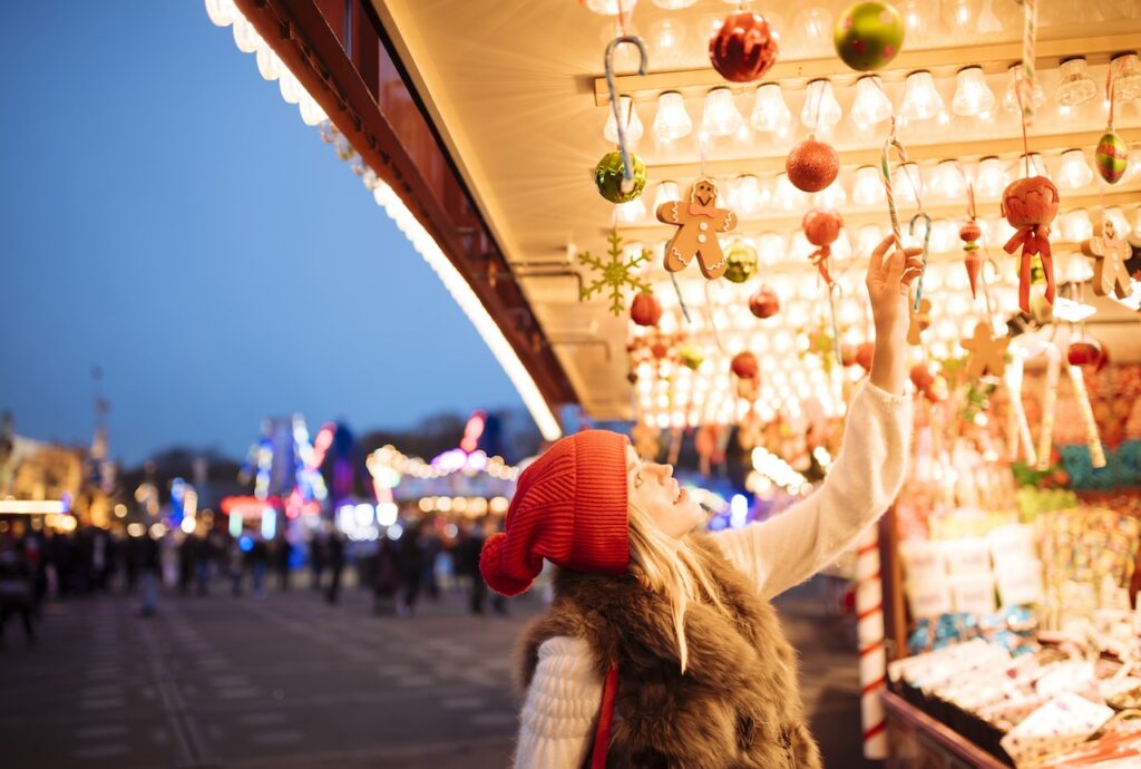 A festive Christmas market stall illuminated at night, with a person in a red hat reaching for hanging ornaments, representing a popular river cruise stop.