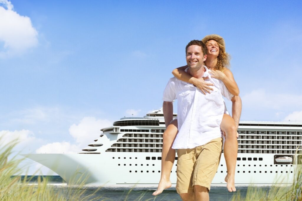 Happy couple on a tropical beach vacation, with a man giving a woman a piggyback ride, set against a backdrop of a large white AmaWaterways cruise ship docked in the distance.
